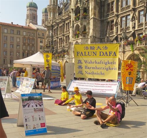 '圖1：二零一九年八月二十四日，法輪功學員在德國慕尼黑瑪琳廣場舉辦活動。圖為民眾跟法輪功學員學煉功法。'