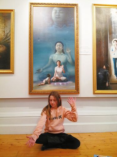 圖5：澳洲女孩何莉坐在《純淨入仙境》畫作前，模仿著畫中女修煉者的姿勢打手印，家人很高興，一家人表示要去煉功點學功。