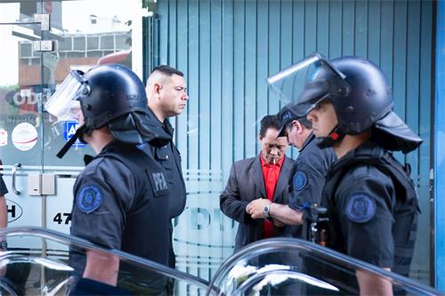 '圖中正在被警察扣押，盤問、身穿紅色體恤深色外套的男子是在二零零四年羅幹到阿根廷時，毆打和平抗議羅幹的法輪功學員的報導，並因此而被阿根廷法院通緝的疑犯。'