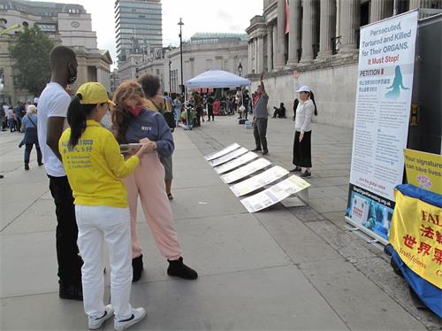 '圖8：二零二零年八月二十二日，在特拉法加廣場，第一次看到法輪功真相的兩位倫敦學生斯卡特（Scalett，正在簽名的女青年）和萬德瑞（Vandre，正定睛觀看法輪功學員煉功男青年）簽名制止迫害。'