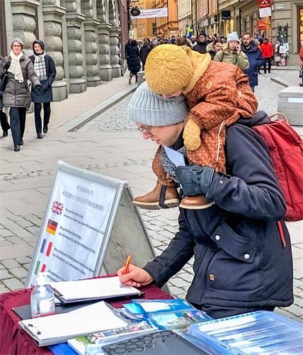 '圖5：這位瑞典人在簽名支持制止迫害法輪功時，騎在爸爸肩上的寶寶認真地注視著爸爸簽名。'