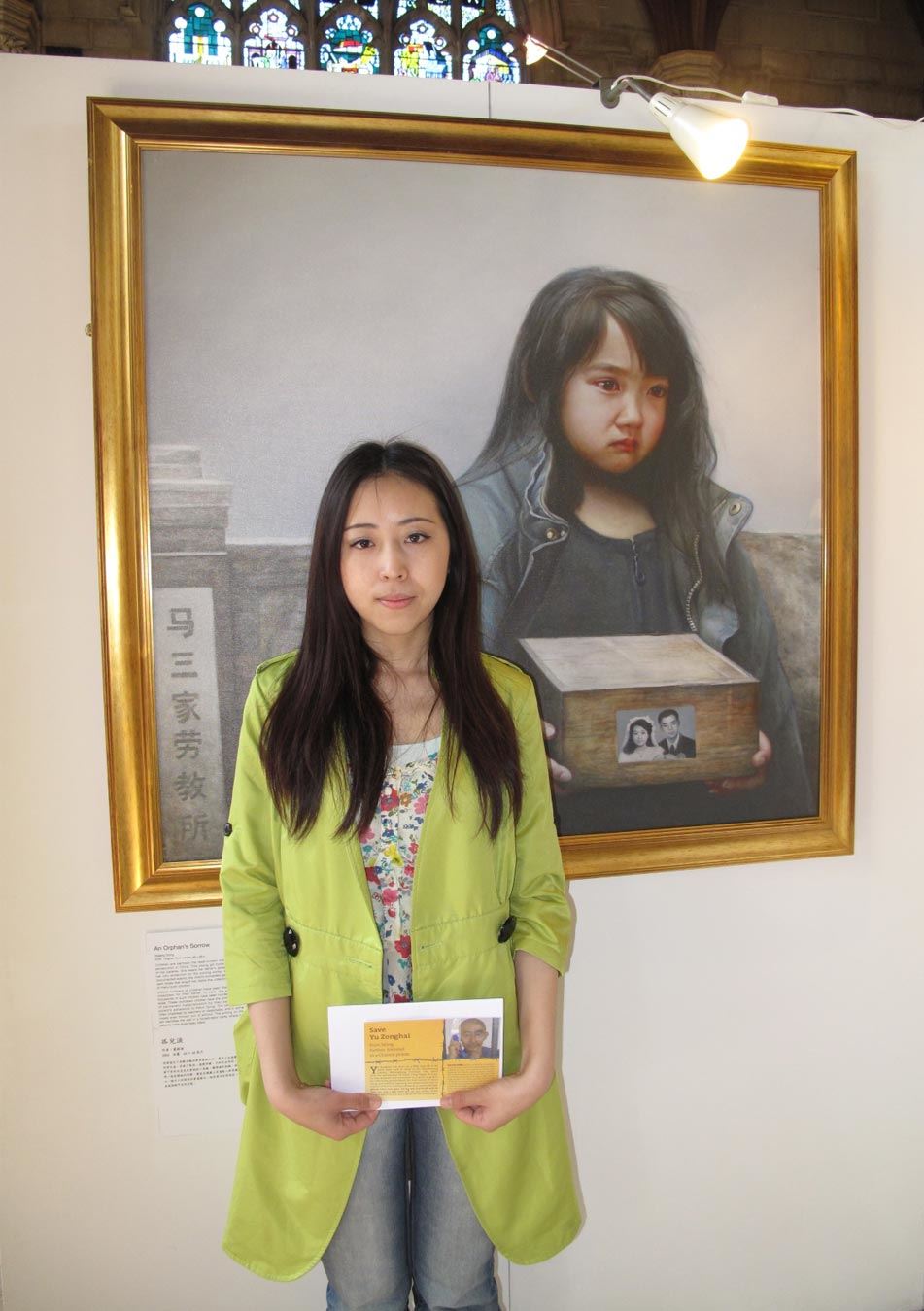 '圖3：二零一三年六月，於銘慧手裏拿著營救父親的明信片站在「真善忍國際美展」畫作《孤兒淚》前。'